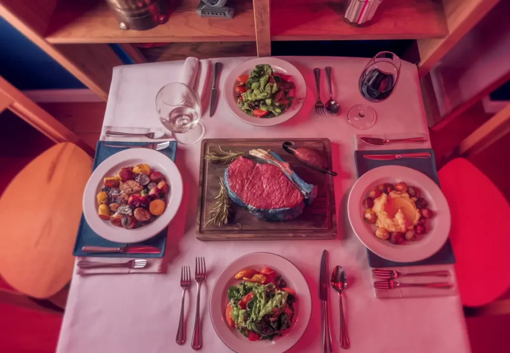 An overhead view of a set table prepared for a blue steak dinner. The centerpiece is a large, rare steak on a wooden board. Surrounding the steak are side dishes of roasted vegetables, mashed potatoes, and fresh salads. Place settings with silverware, empty plates, and wine glasses complete the scene.