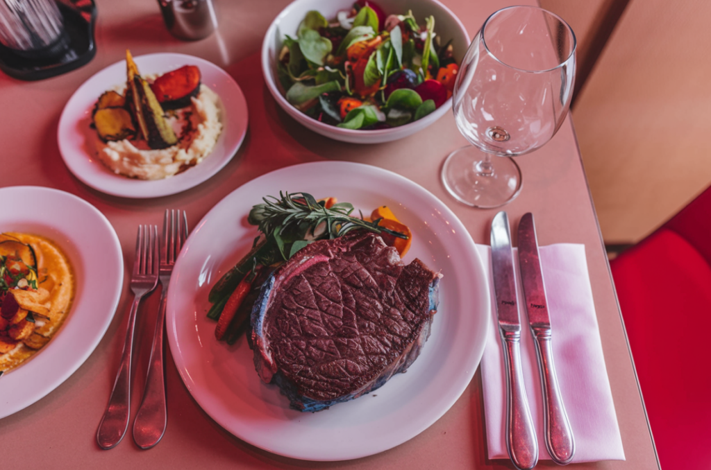 Close-up of a plated blue steak, served with roasted root vegetables and a green salad, accompanied by silverware and a wine glass on a table setting with reddish lighting, highlighting the steak's rare doneness and appealing presentation.