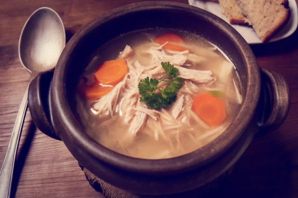 A steaming bowl of chicken soup with shredded chicken, carrots, and parsley garnish.