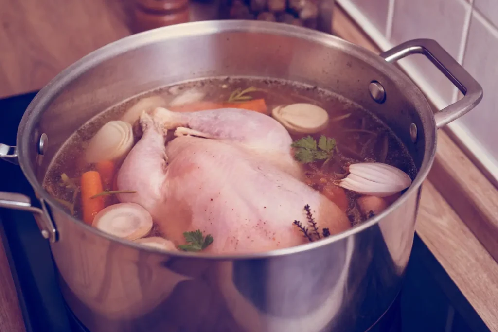 A whole chicken simmering in a stockpot with onions, carrots, and herbs.