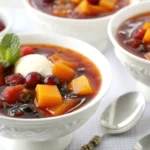 A bowl of Swedish Fruit Soup garnished with mint leaves and a dollop of cream, surrounded by fresh fruits and spoons.